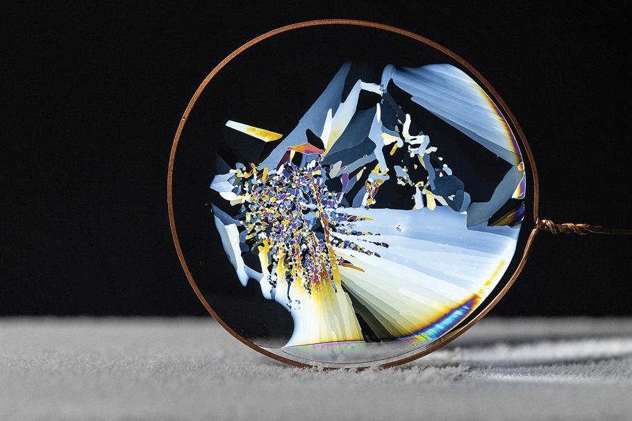 A thin, circular slice of ice is suspended in a round wire frame. Thin white jagged formations collide with one another, producing a rainbow effect evocative of a kaleidoscope.