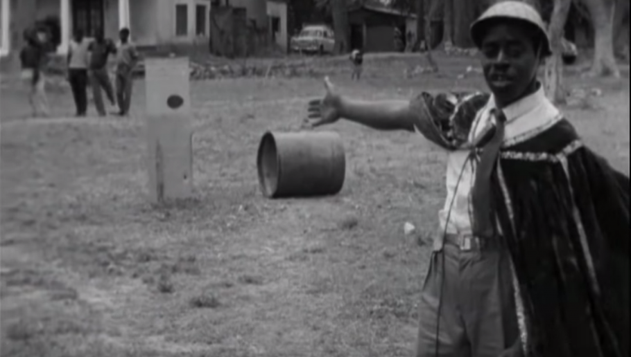 Old photograph on a man pointing toward a small barrel on the ground in a field.