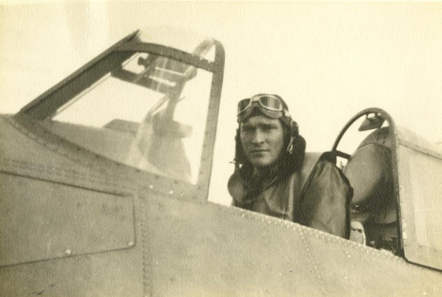A pilot in vintage flight gear, including goggles, sitting in the cockpit of an old aircraft.