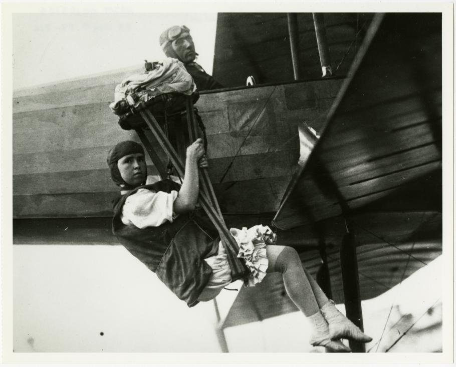A woman hangs from behind the wing of an airplane, looking towards the camera. A man sits in the cockpit, also looking toward the camera.