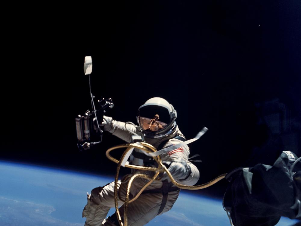An astronaut floating in space. The Earth can be seen below in the bottom half of the frame. The astronaut holds out a long metal rod in one hand.