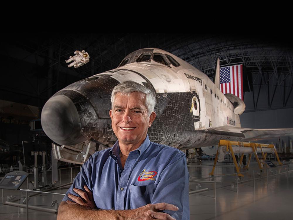 Dennis Jenkins stands in front of space shuttle Discovery—smiling, with his arms folded, and wearing a blue shirt. An American flag is hanging from the ceiling behind Discovery.