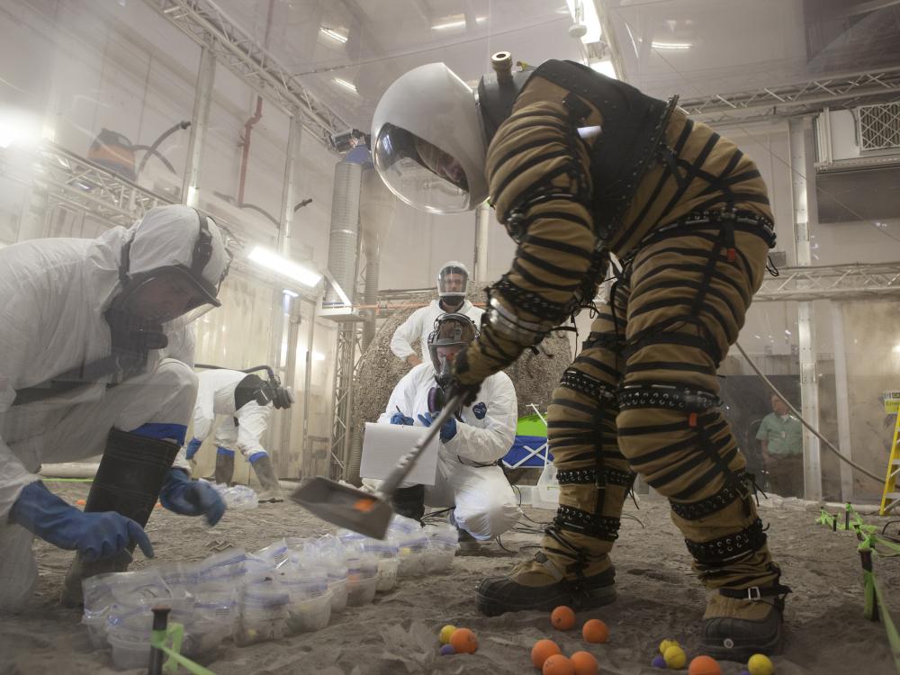 A person outfitted in the new spacesuit uses a tool to pick up balls and place them in bags in front of him. 