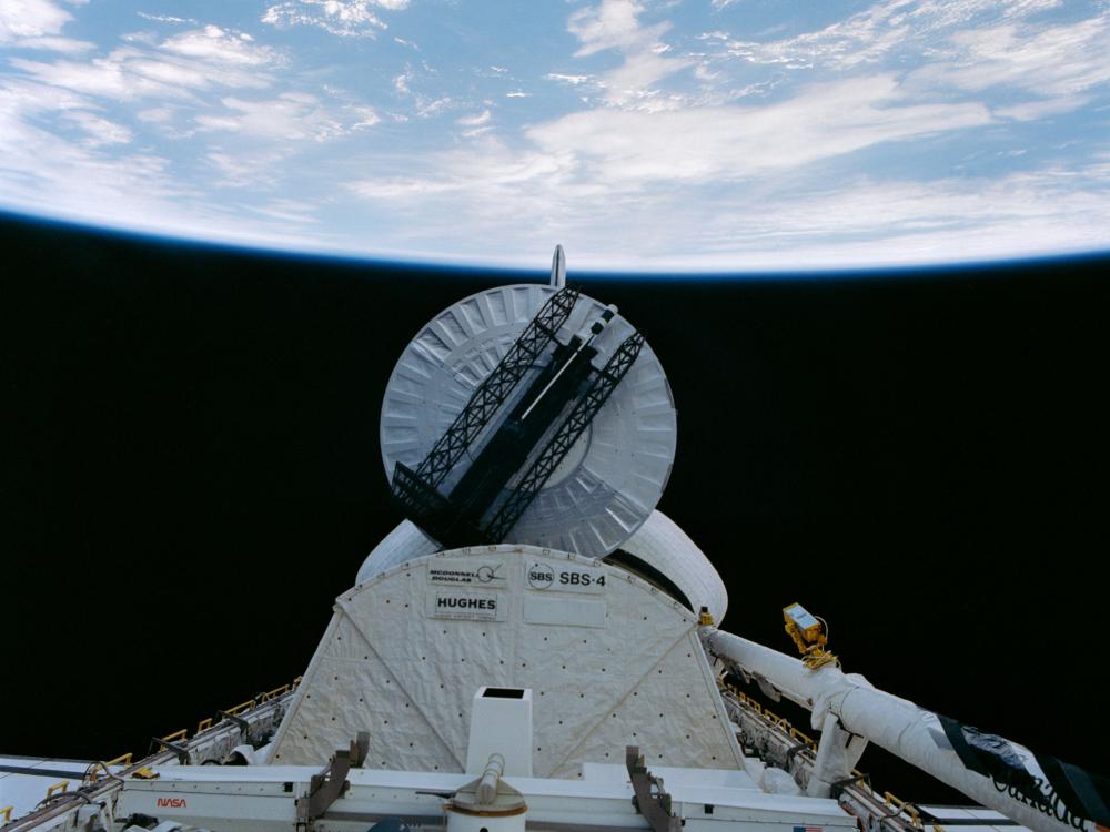 Top view of a satellite acting as payload on a Space Shuttle mission. The satellite has a large, circular top that looks like a dish with an antenna folded above the top.