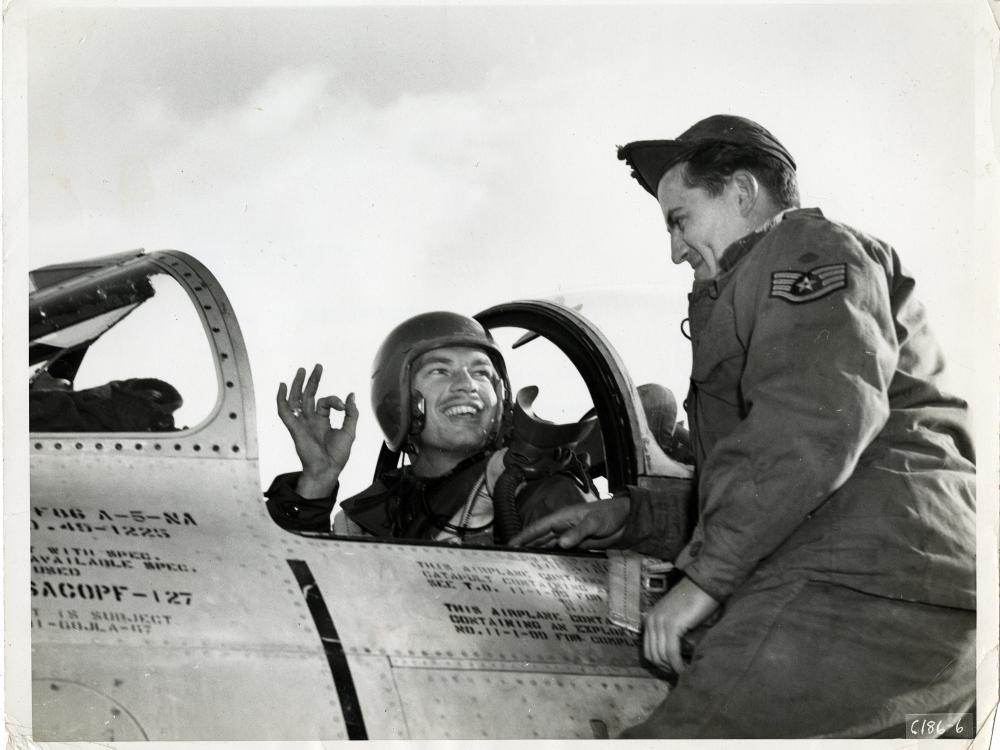 Man in a helmet sits in an aircraft cockpit giving the okay sign with his right hand to a man in a cap and flight suit on his left