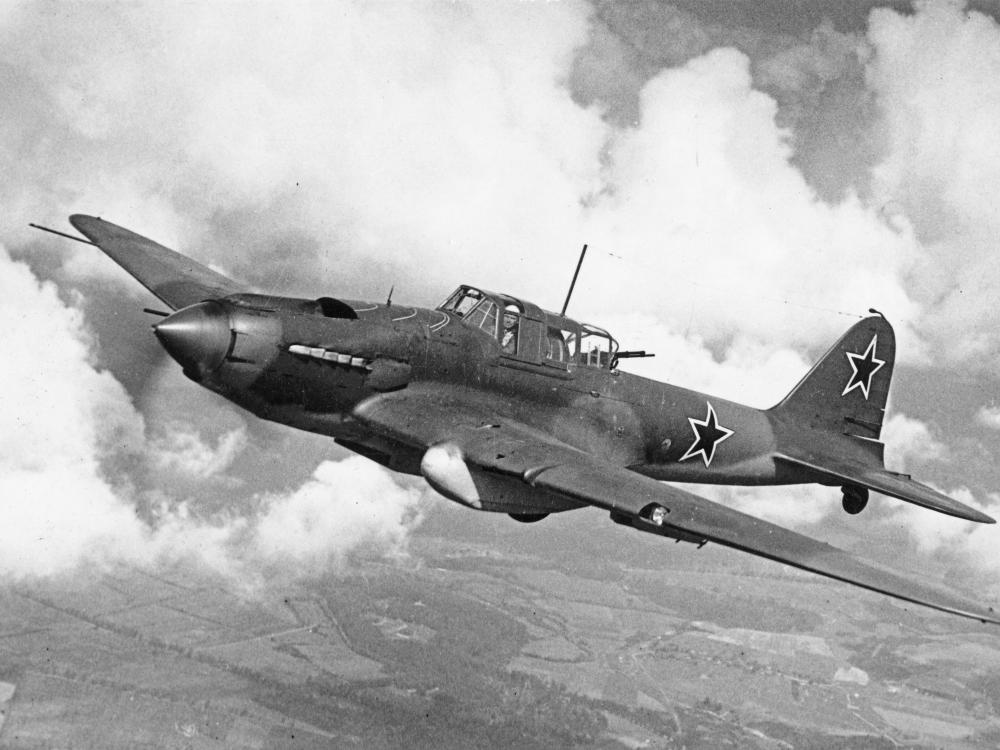 Historical photo of aircraft in flight. Clouds and farmland can be seen in the background. 