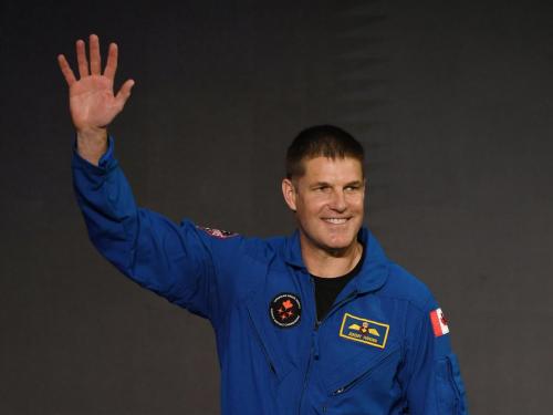 Astronaut Jeremy Hansen, wearing a blue flight suit smiles and waves.