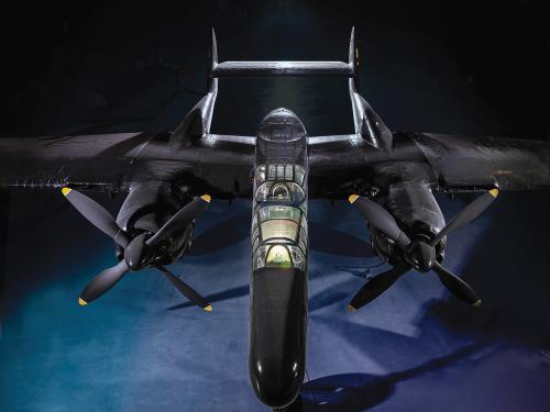 A black, twin-engine airplane is on display inside a dimly lit museum gallery. The airplane has a twin-boom tail and a long nose with a plexiglass-covered forward cockpit.