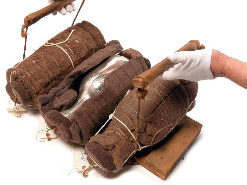 Three flasks wrapped with brown felt and twine lie on top of a hand-made wood tray. A pair of human hands wearing white gloves is holding the tray's two handles.