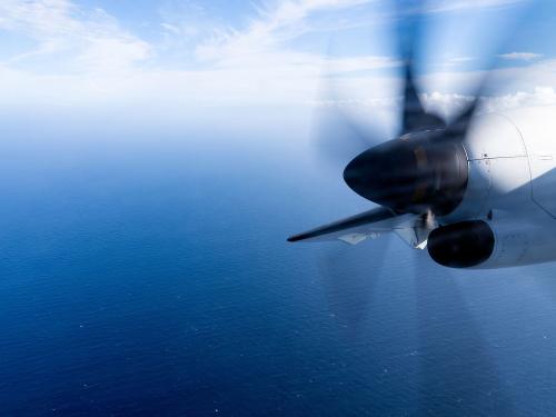 Wing view from a propeller plane flying over a vast ocean with a vibrant blue sky and clouds.