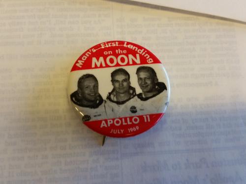 Pin with image of Apollo 11 astronauts in the center and at top and bottom white text against red background. 