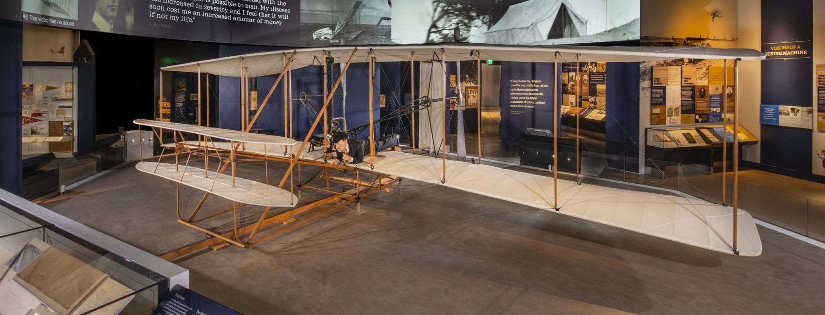 A view of the Wright Flyer in situ, with images and text on the wall behind it. 