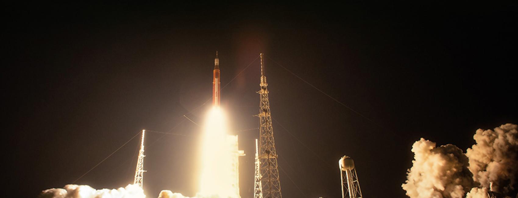 The pitch black night is illuminated by a bright white-hot rocket engine plume, which illuminates clouds of smoke surrounding the launch pad.
