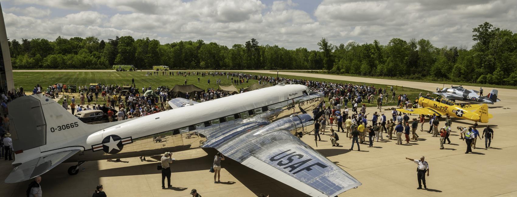 Visitors crowd around historic aircraft outside of the Steven F. Udvar-Hazy Center. In the foreground is a large silver military plane, followed by a smaller yellow one. 