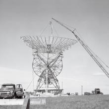 A crane lowers a large radio telescope array on to the ground. The black and white photo sand the style of the truck suggest the photos age.