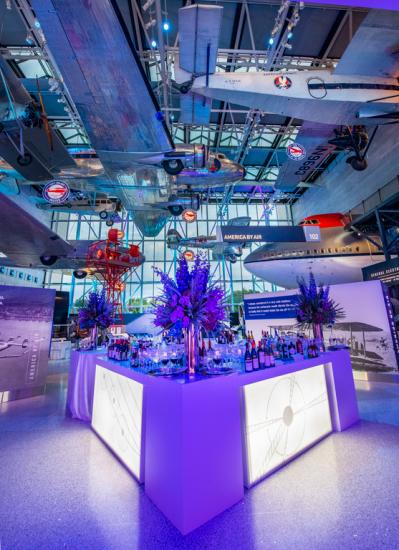 A gallery with planes handing from the ceiling. It's set up for special event with tables with flower arrangements on them on the floor. 